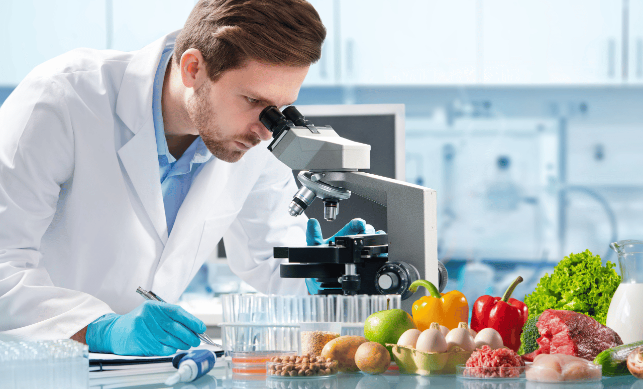 Food safety scientist examining samples under a microscope with fresh produce, meat, dairy, and grains in a laboratory setting – Phoenix Food Consultancy